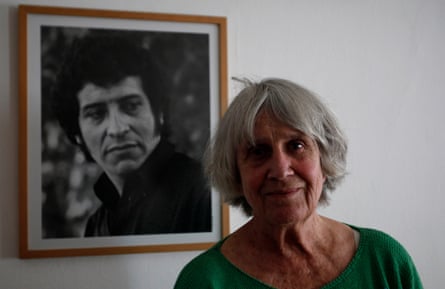 Joan Jara smiling in front of a framed photo of Víctor Jara on a wall