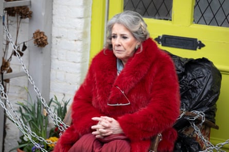 A woman in a red coat sits in front of her yellow front door, chained to the porch