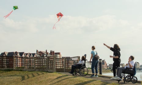 Two people in wheel chairs either side of two people standing. They are flying two kites and on a path near water