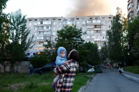 A woman carries a toddler as smoke rises above a Kyiv multi-storey building after Russia attacked, hitting apartment buildings