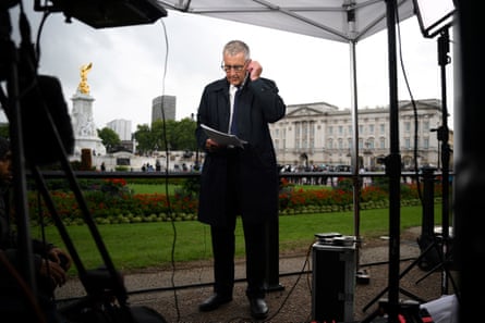 Reading notes with his left-hand to his ear, suggesting he’s in communication with his producers, Dermot Murnaghan stands surrounded by lights and under a canopy, with Buckingham Palace in the distance