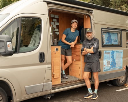 Woman in shorts and T-shirt standing inside grey camper van, next to man in shorts and T-shirt bearing charity logos.