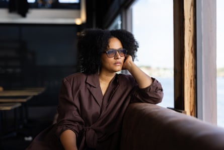 A woman wearing an oversized brown suit jacket and sunglasses sits on a couch with water seen through the window behind her.