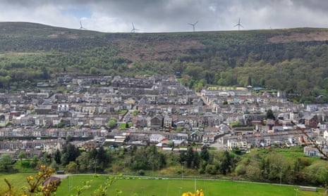 A view of a town nestled in a green valley, seen from above, with wind turbines in teh distance on the far ridge of the valley