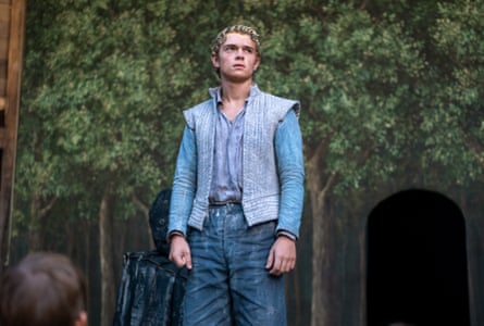 A young man with bright blond hair looks to the side while standing on stage in a film still.