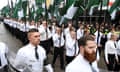 A long double line of all white people in white shirts, black pants, and black ties, holding up dark green, white and black flags, march and appear to chant on a wet ur