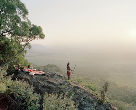A champagne picnic laid out on a hill in the Maasai Mara, Kenya, with a Masaai tribesman looking out over the valley.