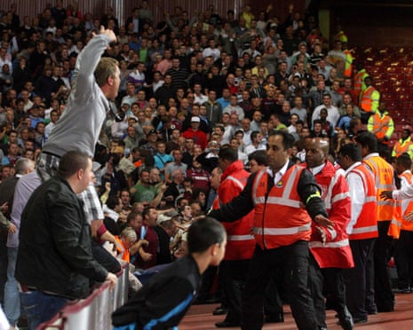 A fan stands on a fence as stewards in orange bibs gesture for fans to stay in a stand