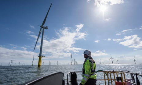 A worker in a hard hat at the bow of a ship looks up at the nearest of dozens of huge wind turbines arranged in rows in the sea