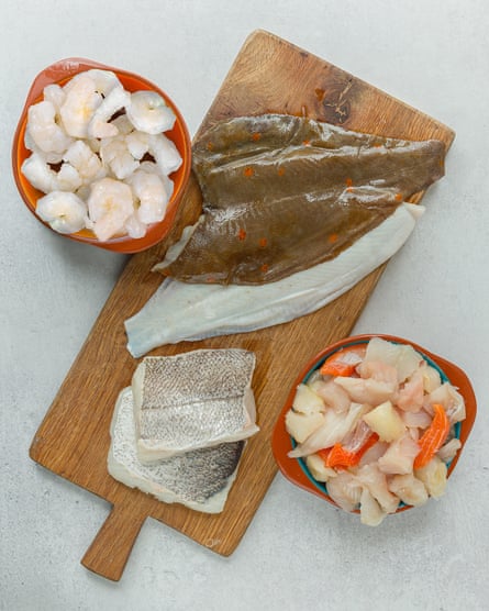 A flat fish and other fillets on a wooden board with a bowl of prawns and another of pieces of fish