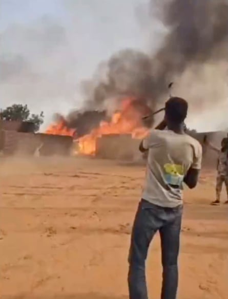 A grainy image showing two men standing a few metres from a burning building, one holding an assault rifle, the other apparently holding up a phone