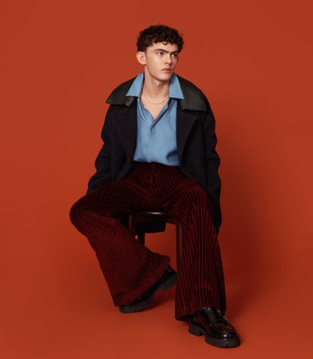 Joe Locke, with short dark curly hair, wearing a blue shirt, a dark coat, dark red flared trousers and shiny black shoes, and sitting in front of a dark orange background