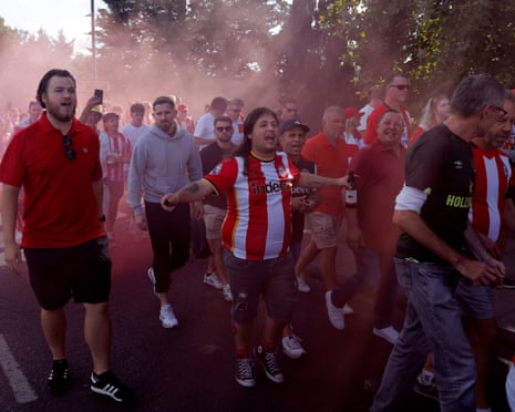 Brentford football fans walk to the stadium through pink smoke from a flare