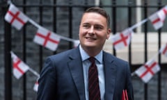 Wes Streeting leaves Downing Street after attending a cabinet meeting: he is seen in a head and shoulders view in front of black iron railings on which England flags are hung as bunting. He is a young man with short fair hair and is smiling; he wears a dark blue jacket, pale blue shirt and dark tie with red horizontal stripes, and holds a red folder under his arm.