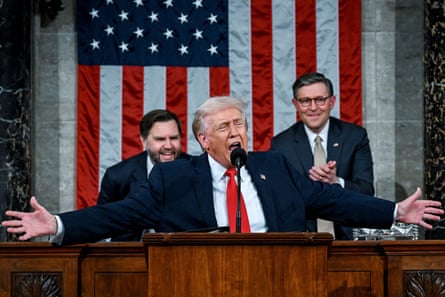 Trump speaks behind a podium with his arms outstretched. Behind him, JD Vance and Mike Johnson applaud