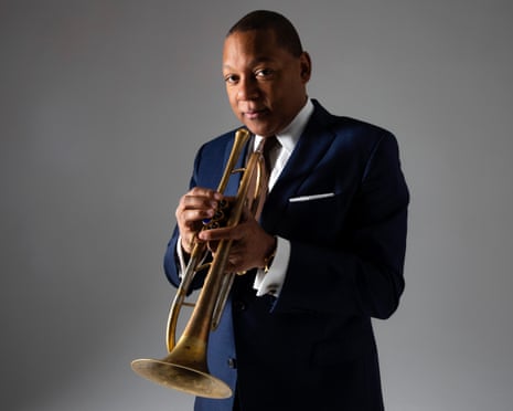 Wynton Marsalis in a navy suit holds a trumpet while looking at the camera against a grey background