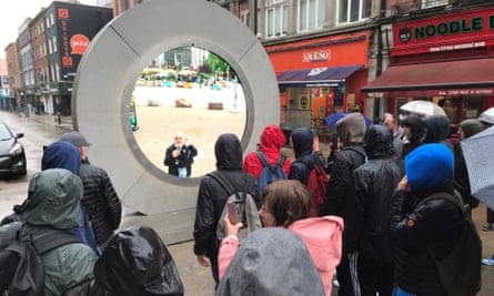 People in a Dublin street stand looking at the circular portal, in which a bald-headed man at the New York side stands looking back