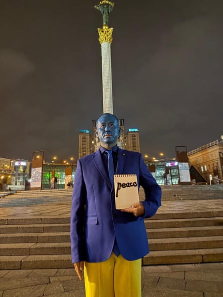 The artist Du Yinghong in Ukraine, standing in front of a monument, face painted blue and yellow, holding a sign that says Peace