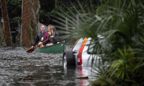 Two women and a man in a green canoe ppaddle through a flooded street, past a silver crossover car that is mostly submerged. The water looks dark and cloudy. 
