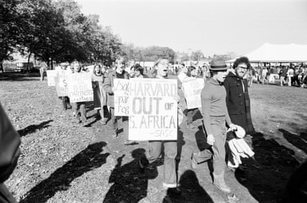 people walk on a field with signs, including one saying ‘harvard out of africa’, in a black and white photo