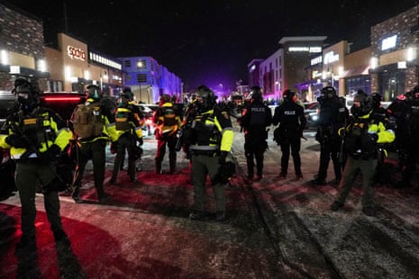 A line of law enforcement officers in riot gear and high-vis clothing stand across a wintery street that is backlit by red-and-blue police lights.