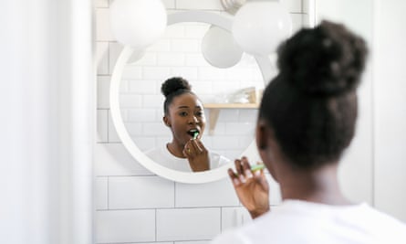 Молода афроамериканка чистить зуби, стоячи перед дзеркалом у світлій ванній кімнаті.