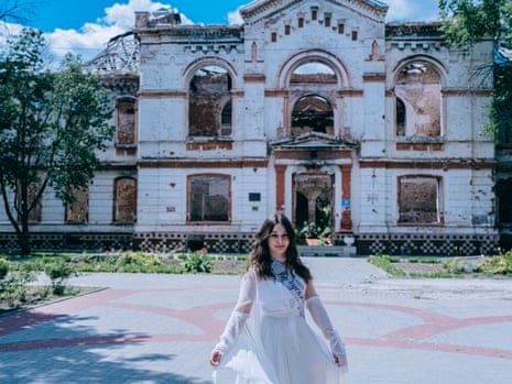 A teenage girl in a white dress stands in front of the gutted shell of a public building