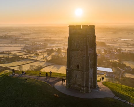 Glastonbury Tor at sunrise, with people walking nearby and misty fields below