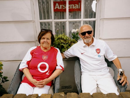 A woman in red Arsenal top sits next to a man with a white beard and sunglasses wearing a white polo shirt and white trousers in front of. a white house with an Arsenal scarf in the window