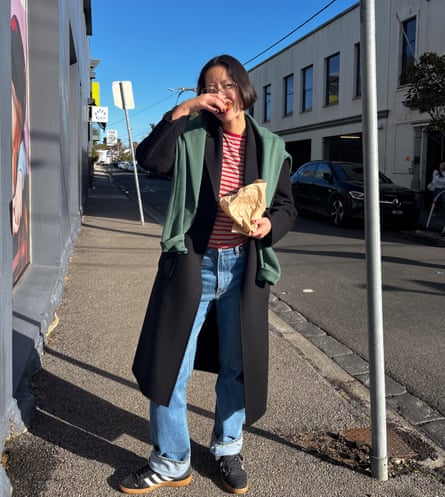 Maggie wears a green hoodie slung over her black coat. She’s also wearing blue jeans and a red striped top.
