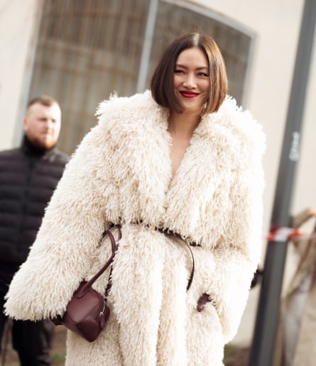 A young woman with a dark bob and red lipstick, wearing a shaggy white coat with a brown belt, bag and gloves