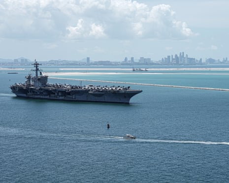 Aircraft carrier in water with city in the distance.