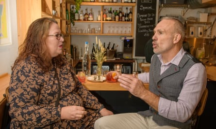 A woman with long red wavy hair, glasses and red lipstick, wearing a brown and black patterned dress, and a man with short grey hair, wearing a lilac shirt, a grey herringbone waistcoat and beige chinos, sitting at a restaurant table talking