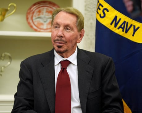 Larry Ellison sits in the Oval Office in front of a US Navy flag; he wears a dark jacket, white shirt and broad red tie and has a neatly-trimmed beard and fair hair. 