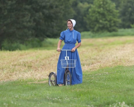 A woman in a field with trees in background in Amish costume (blue dress and white bonnet) holding on to a small bicycle with a basket