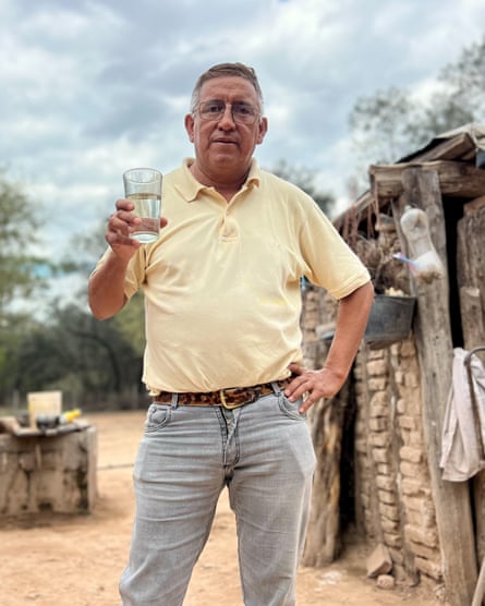 An older man in front of a hut holds out a glass of water