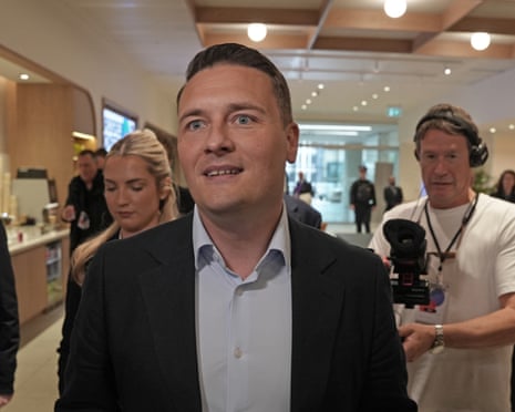 Streeting in a dark suit walks through a modern interior surrounded by media and cameras
