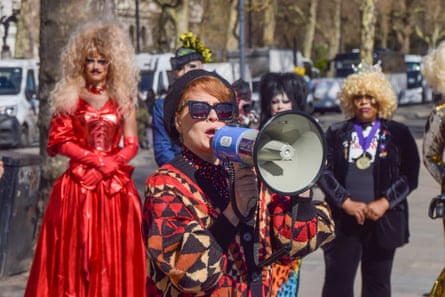 Ana Martronic speaking into a megaphone, wearing a patterned jacket, and black hat and sunglasses, with a group of drag queen behind her