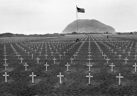Rows of crosses in a field while a US flag flies in front of a large mountain.