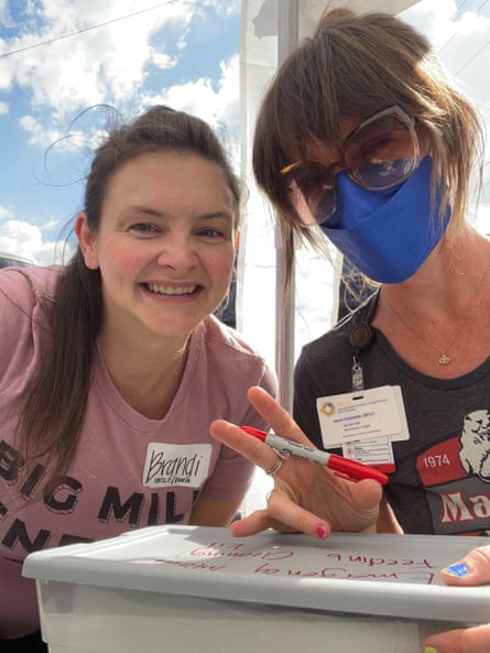One woman in a pink shirt smiles next to another woman wearing a sunglasses and a blue face mask, who is in the middle of labeling a plastic storage bin