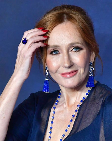 JK Rowling, with long earrings, a long beaded necklace, a ring and painted fingernails, smiles as she puts her hand to her head