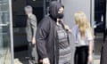Holly LeGresley walks out of a doorway wearing a black hooded top with the hood up and a black face mask, and is using a walking stick