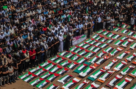 Palestinian flags cover the bodies of the deceased on the ground as a large crowd of people stand nearby in prayer.