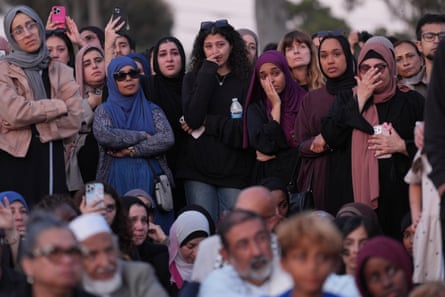 A crowd of mourners, many wearing hijabs and prayer hats, gathered outside.