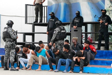 Masked police officers carrying weapons guard a group of handcuffed young men, many of them without shoes, sitting on a set of blue steps.