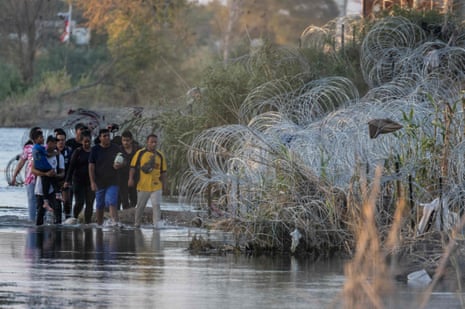 A group of people walk along a shallow river with razor wire heaped on its banks.
