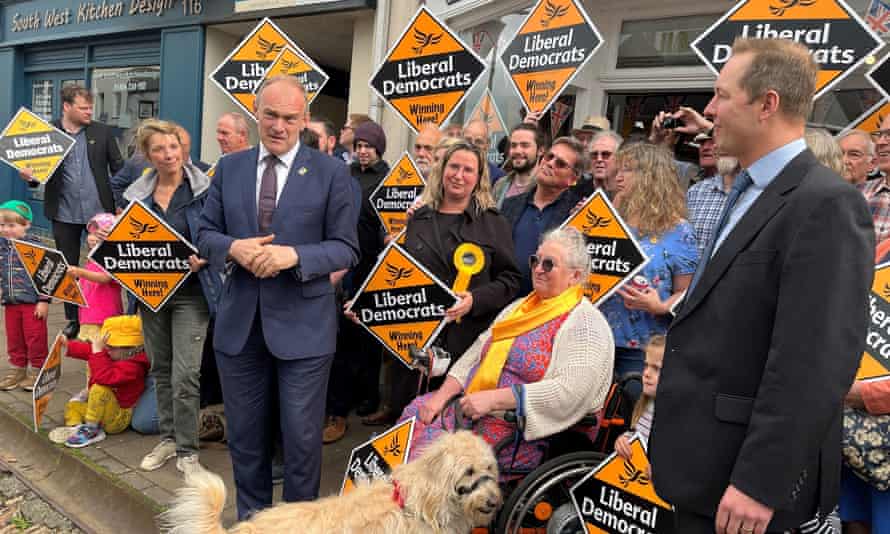 Two men standing next to a woman using a wheelchair with a dog, with a backdrop of people holding up orange Lib Dem signs