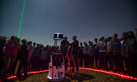 Un cercle d'enfants plus âgés entoure un grand télescope tandis que l'un d'entre eux se tient debout sur un escabeau pour regarder à travers. 
