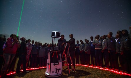 A circle of older children surround a large telescope as one stands on a stepladder to look through it