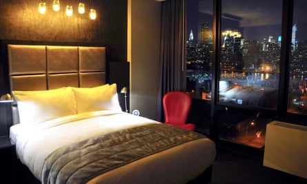 Illuminated lightbulds hang above a double bed in a room at the Z NYC Hotel in New York City. Out of the room windows in the NYC skyline at night.